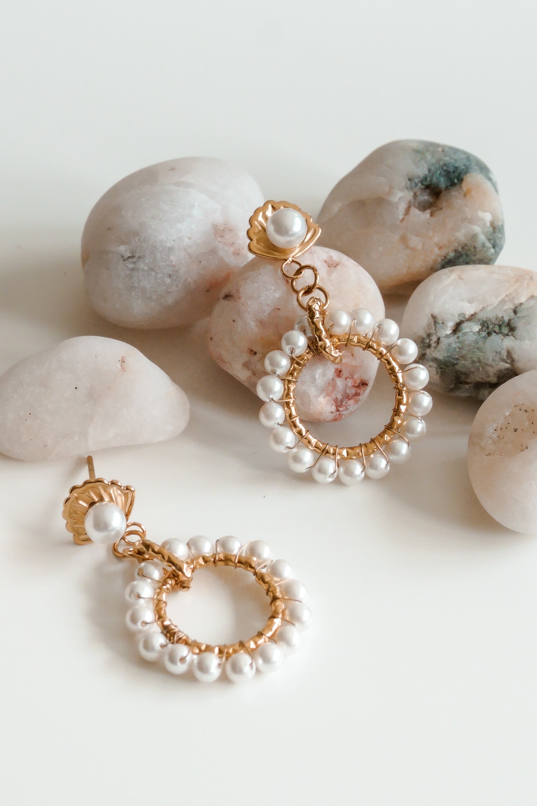 Mazie auskari - Pērļu dārzs by Como Jewelry - zelta riņķveida auskari ar perlamutra pērlēm - ir izvietoti uz baltas virsmas starp gaišiem akmentiņiem, izceļot šo eleganto pāri.