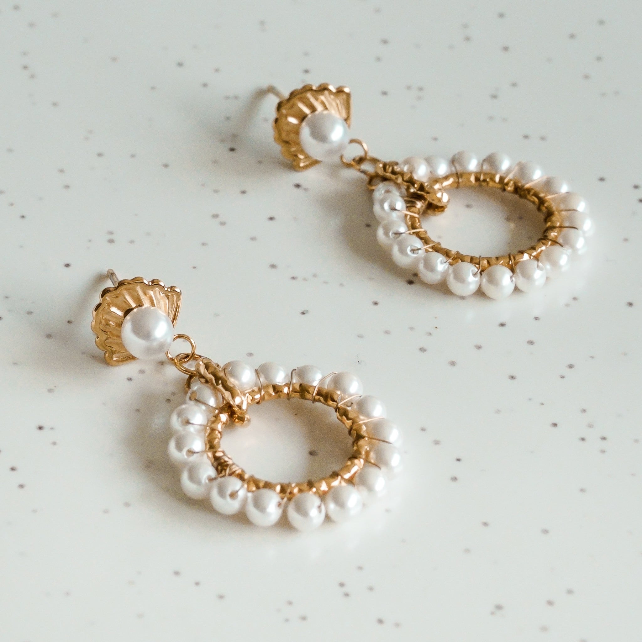 Mazie auskari - Pērļu dārzs no Como Jewelry ir zelta un pērļu auskari ar apaļiem riņķīšiem, ko rotā maigas pērles, kas izvietotas uz baltas punktētas virsmas. Šie elegantie auskari jebkuram tēlam piešķir mūžīgu stilu.