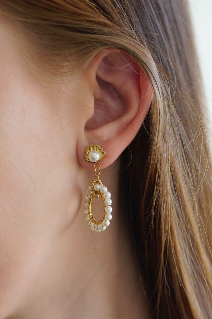 Sievietes auss tuvplānā ar Mazie auskari - Pērļu dārzs by Como Jewelry: zelta auskari ar nelielu pērļu uzgaļu un ovālu cilpu, kas akcentēta ar perlamutra pērlēm. Viņas brūnie mati ir aizķērušies aiz auss.