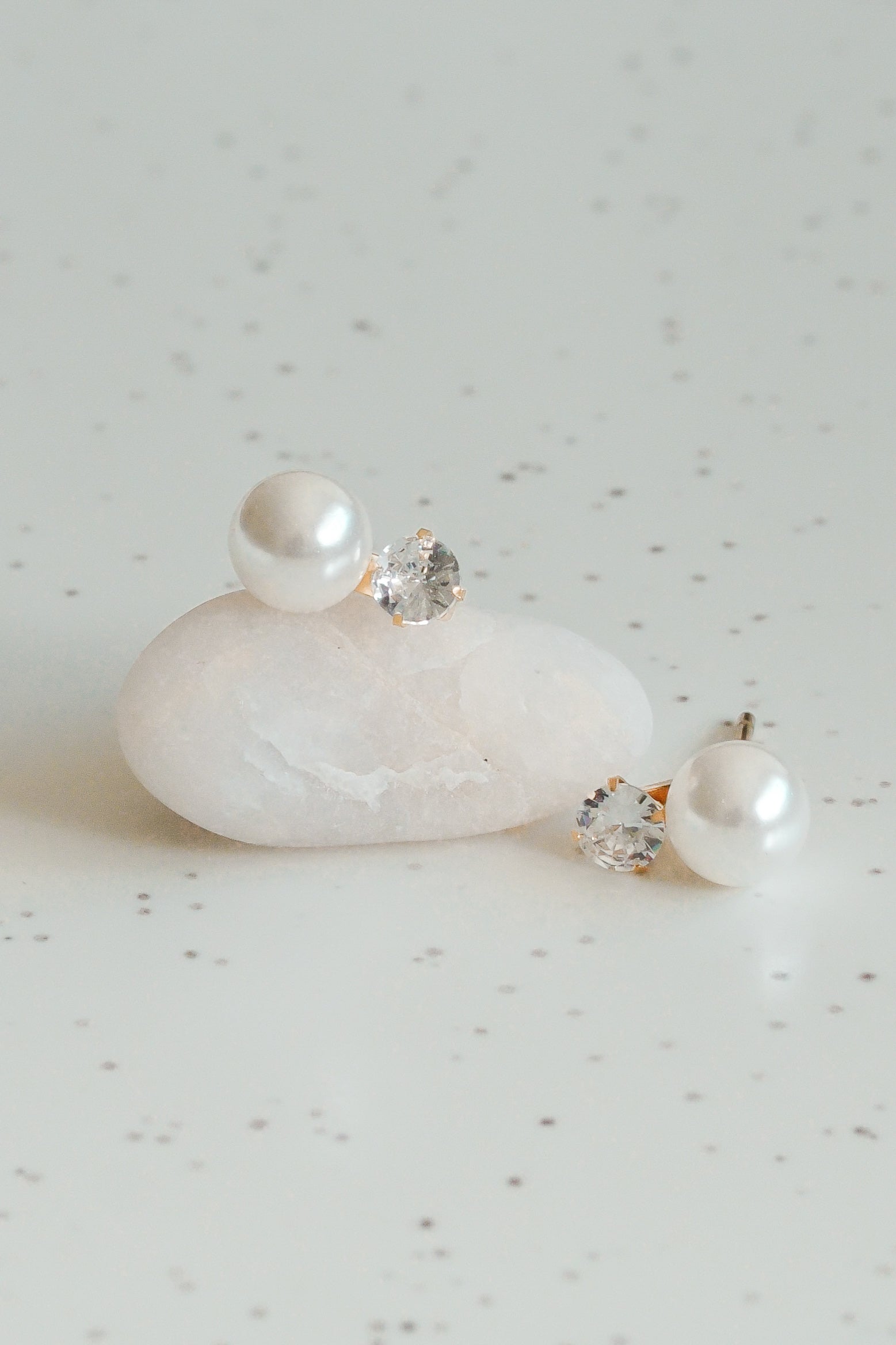 Mazie auskari - Klusā gaisma by Como Jewelry: zeltīti minimālisma auskari ar apaļām baltām pērlēm un caurspīdīgiem dārgakmeņiem, kas izvietoti uz gluda balta akmens ar gaišu, plankumainu fonu.