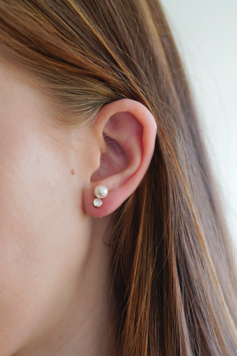 Tuvplāns personas ausij, kurai ir Como Jewelry Mazie auskari - Klusā gaisma: zeltīts minimālisma stila pērļu imitācijas auskari virs mazas apaļas dimanta formas nagliņas, ar taisniem, gaiši brūniem matiem, kas novietoti aiz auss.