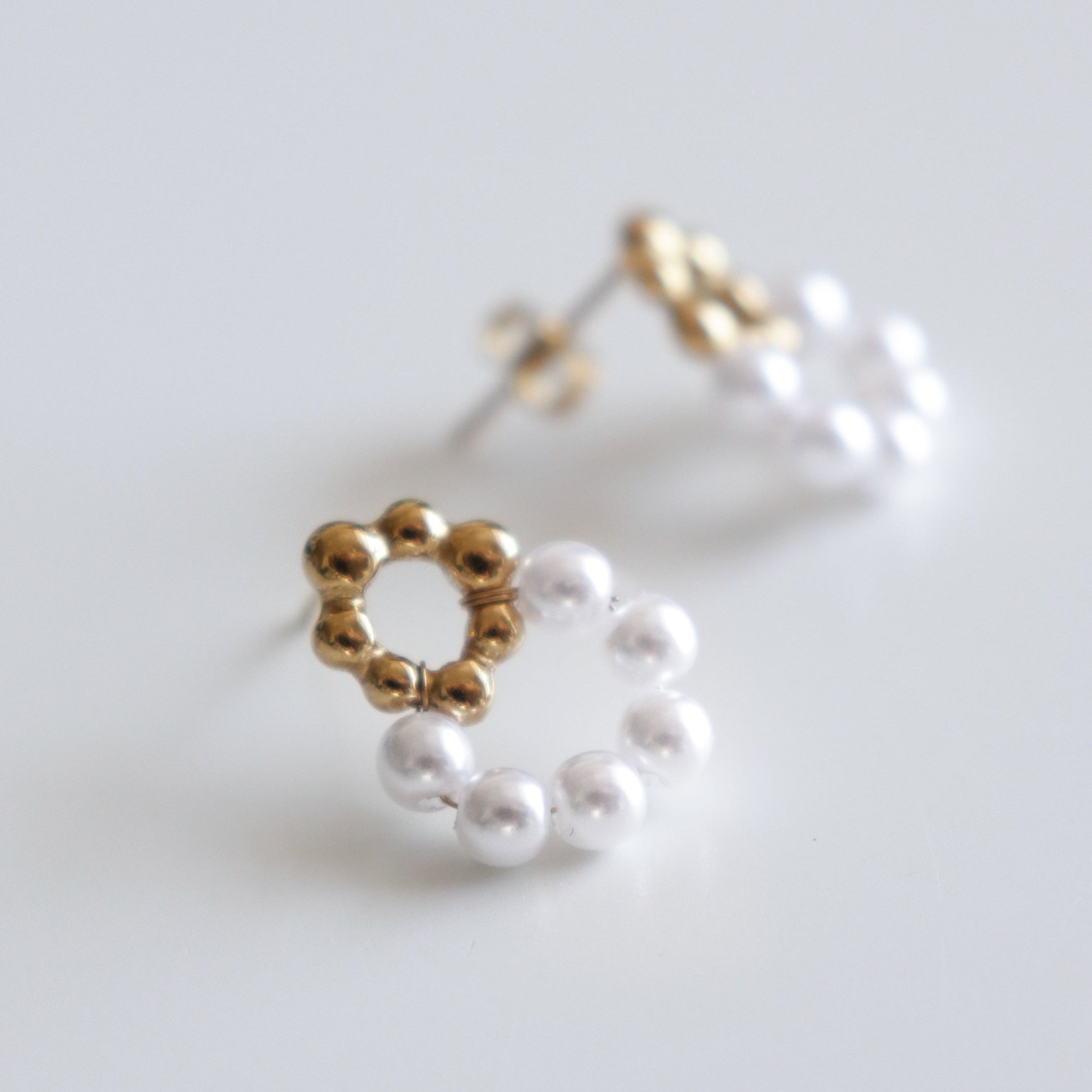 Mazie auskari - Pērļu ziediņi by Como Jewelry: apzeltīti nerūsējošā tērauda auskari ar mazām zelta pērlītēm, kas veido ziedu, un baltām pērlēm līdzīgām pērlītēm aplī, attēlotas uz balta fona.