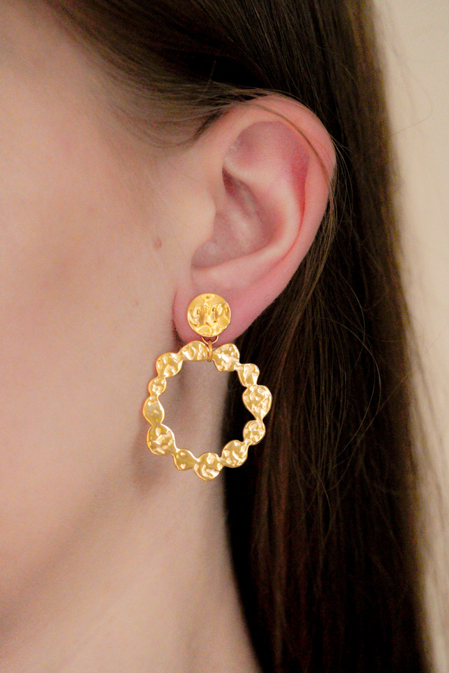 Tuvplāns cilvēka ausij, kam ir Como Jewelry "Auskari - Saules Rats", lieli tekstūras auskari ar zelta pārklājumu, ar apļveida dizainu un apaļu tapu. Personai ir taisni brūni mati, kas aizbīdīti aiz auss.