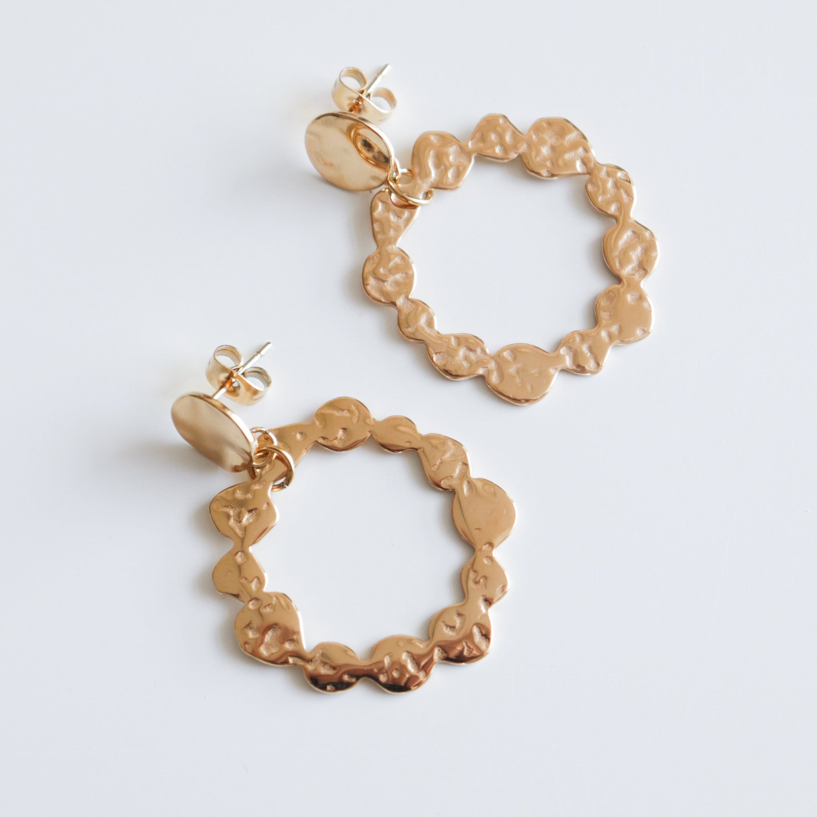 Auskari - Saules Rats by Como Jewelry: zelta pārklājuma juveliertērauda auskari ar nelīdzenām, viļņotām malām un kaltu tekstūru, attēloti uz balta fona.