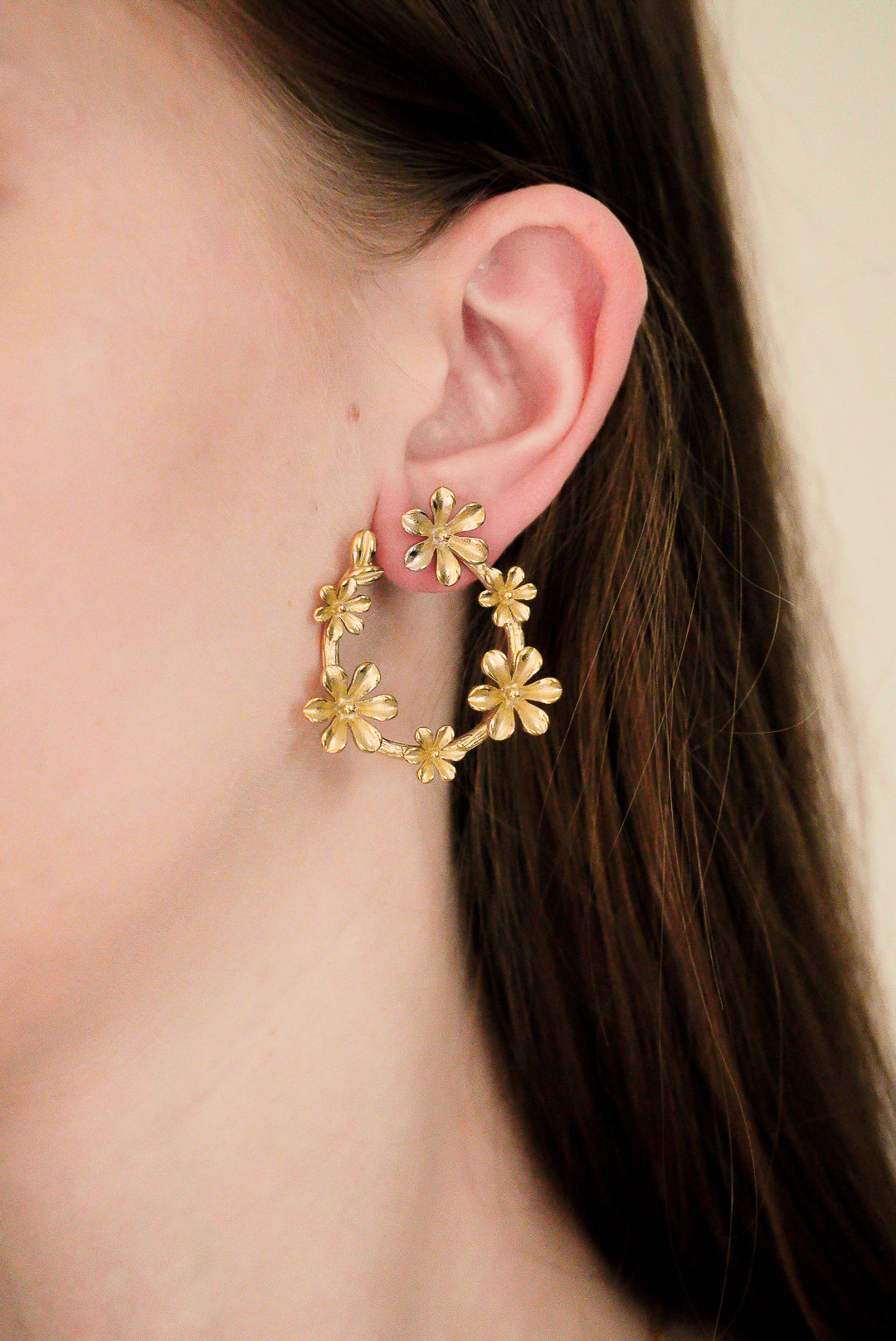 Tuvplāns sievietes ausij, kurai uzliktas Como Jewelry "Auskari - Rokoko" - lielas, apzeltītas cilpas ar sarežģītu ziedu rakstu. Unikālie auskari izceļas uz viņas garo brūno matu fona, kas daļēji redzams attēlā.