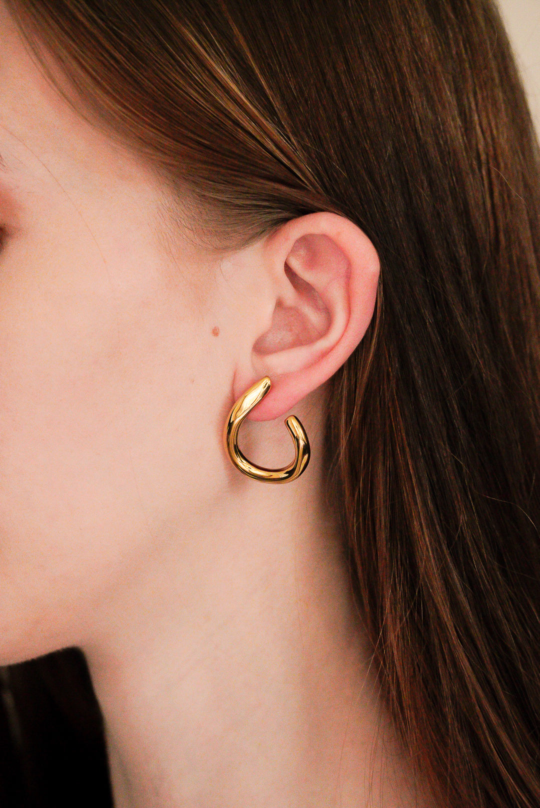 Tuvplāns sievietes ausij, kurai ir Como Jewelry Auskari - Plūsma, biezi zelta abstraktas formas auskari. Viņas taisnie gaiši brūnie mati ir aizķērušies aiz auss, un gaišā āda kontrastē ar neitrālo fonu.