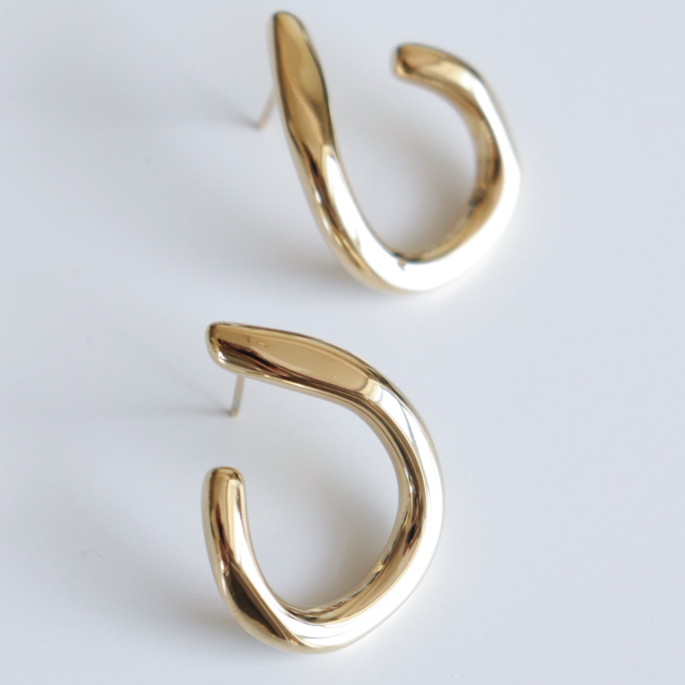 Auskari - Plūsma no Como Jewelry ir spīdīgi zelta auskari ar modernu, izliektu dizainu uz balta fona.