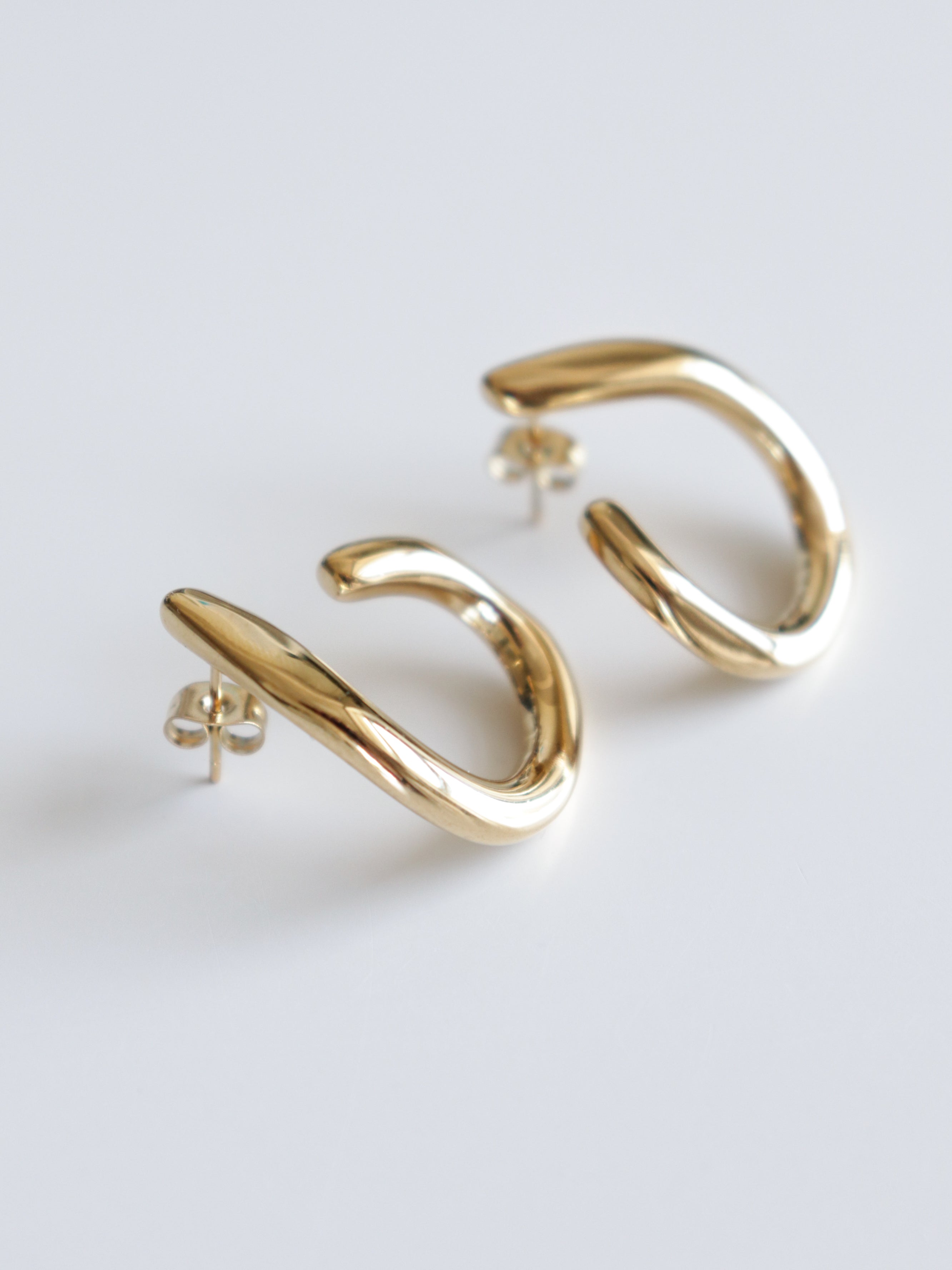 Como Jewelry-Plūsma veidots pāris spīdīgu, zeltītu, izliektu hoop auskaru auskaru, kas izgatavoti no ūdensizturīgi juveliertēraudiem ar push-back aizdari, kas redzami uz balta fona.