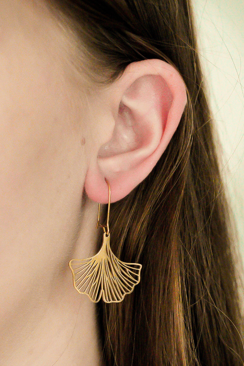 Sievietes auss, kas rotāta ar Como Jewelry Auskari - Ginkgo, tuvplānā, viņas brūnie mati aizbīdīti aiz auss.