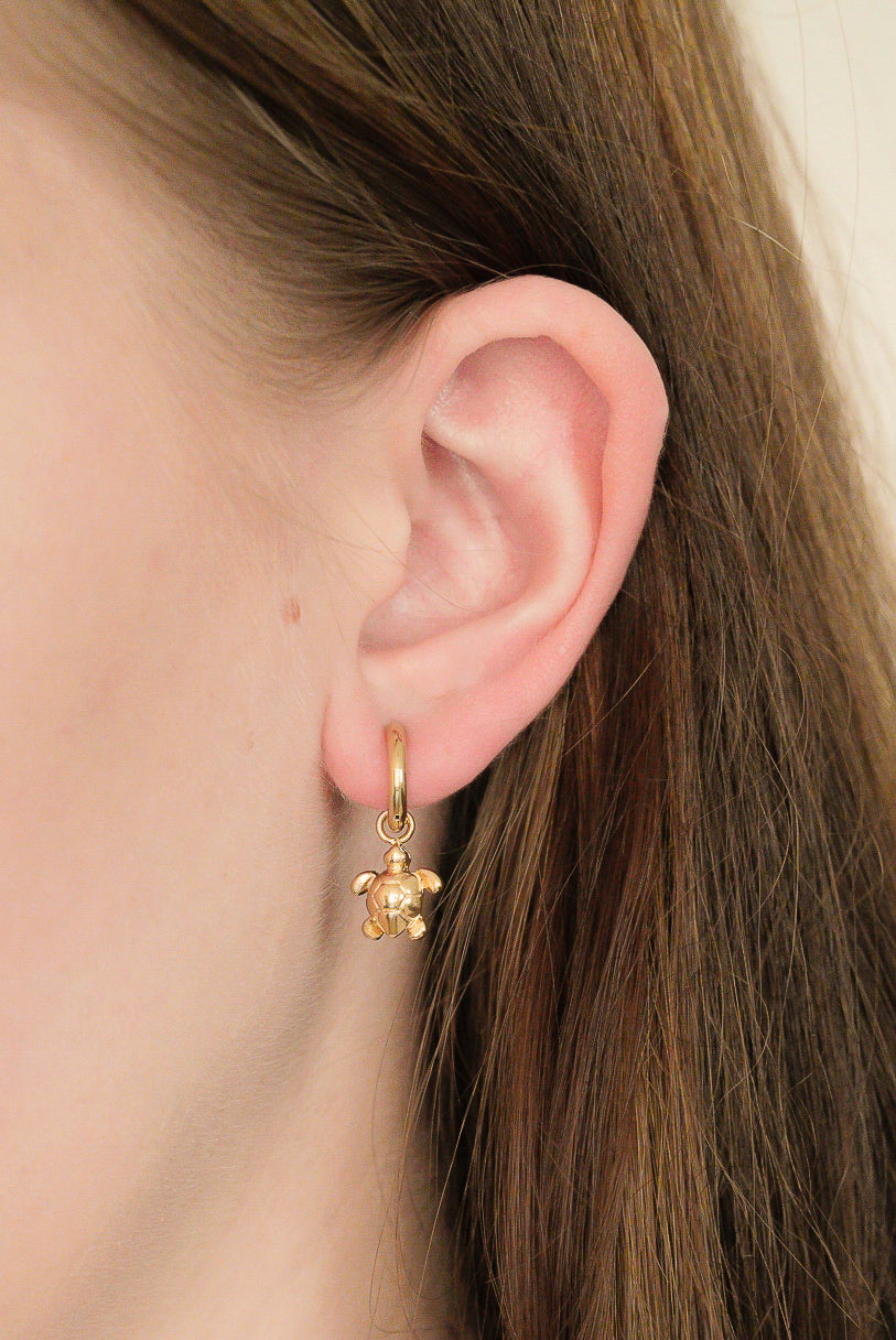 Close-up of a woman’s ear with long brown hair, wearing Auskari – Miers earrings featuring a small teddy bear charm.