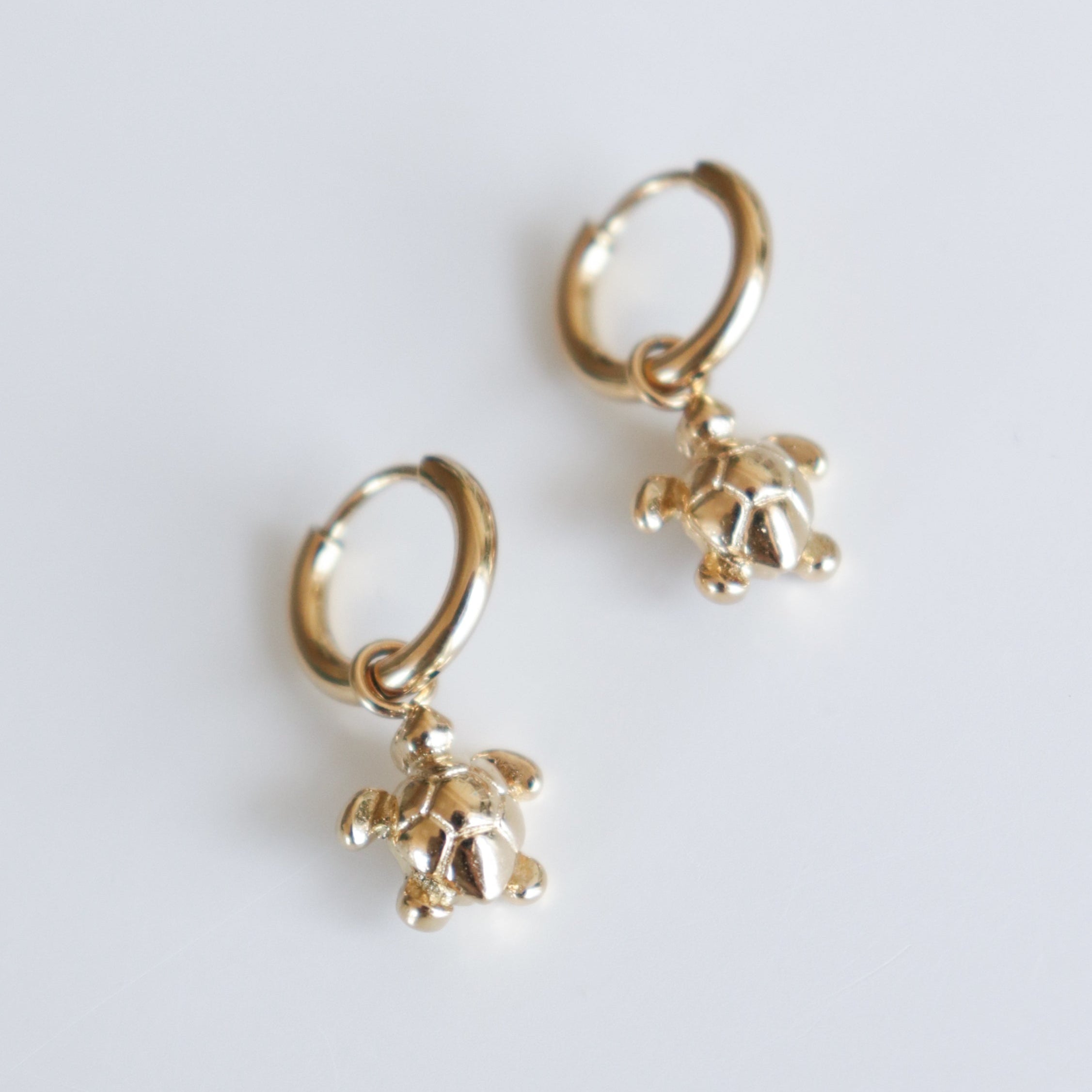 A pair of Auskari - Miers, each featuring delicate bruņurupuču piekariņi, is displayed on a white background.