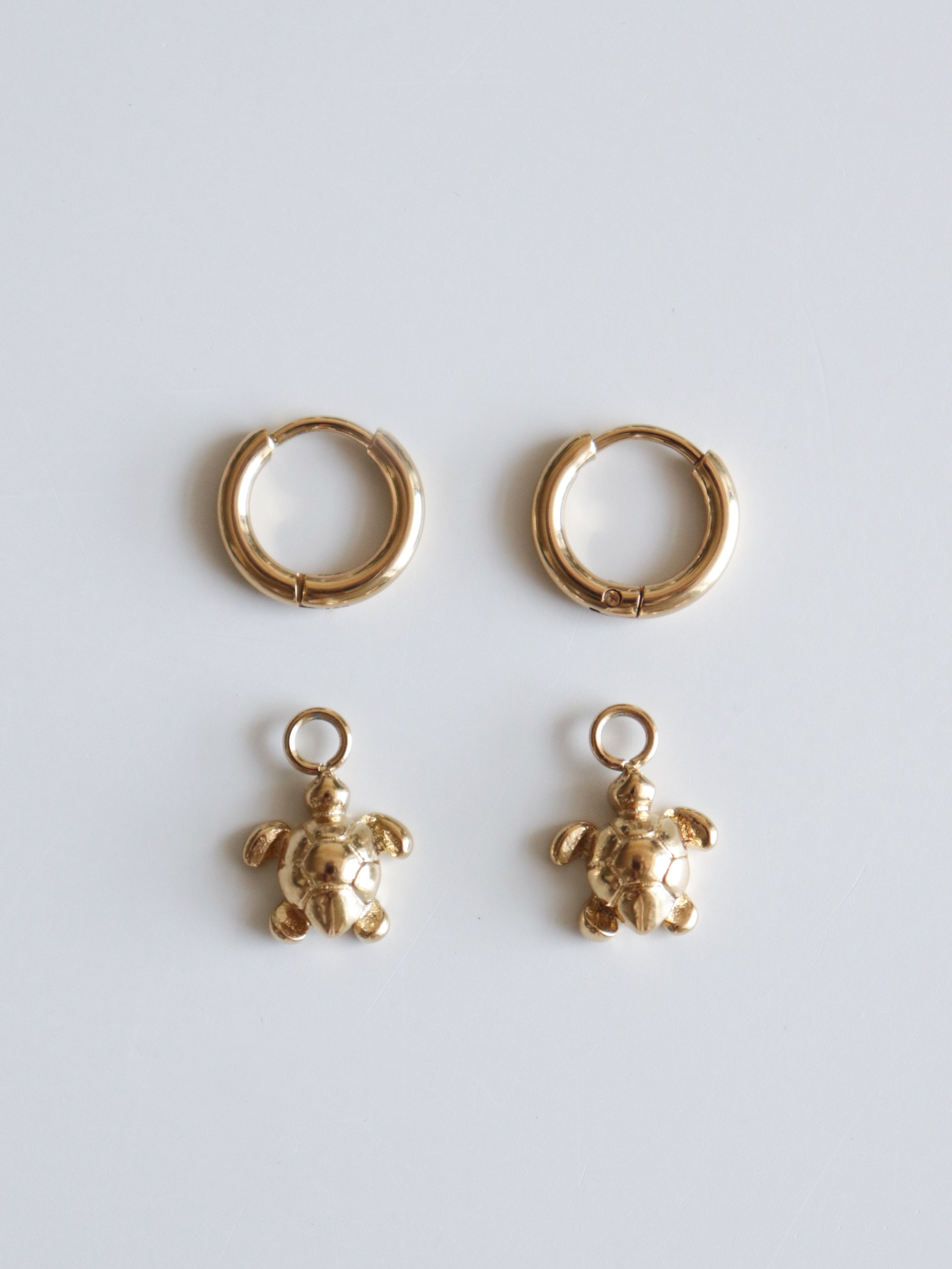 Two zeltīti Auskari - Miers and two gold turtle charms are displayed on a plain white surface, with the charms placed below the earrings, showing they can be combined to create turtle-themed earrings.