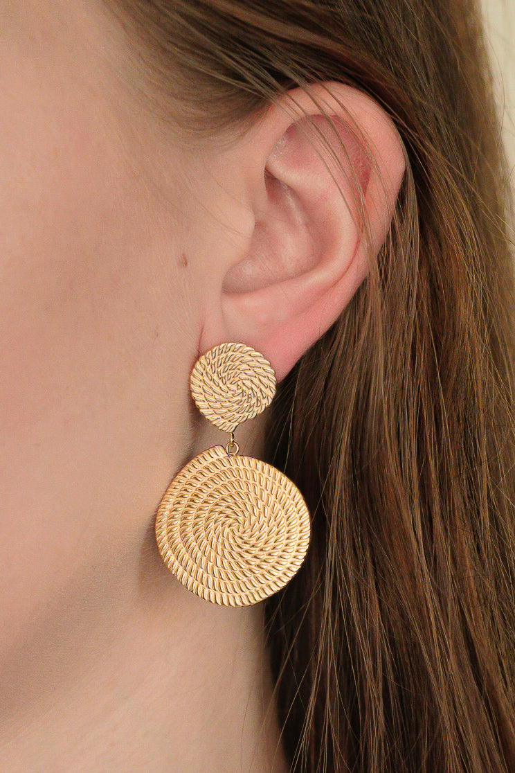 Close-up of a womans ear wearing Auskari - Grācija: gold textured dangle earrings with a small circle top and larger spiral below, crafted from zelta pārklājuma juveliertērauds. Her brown hair is tucked behind her ear.