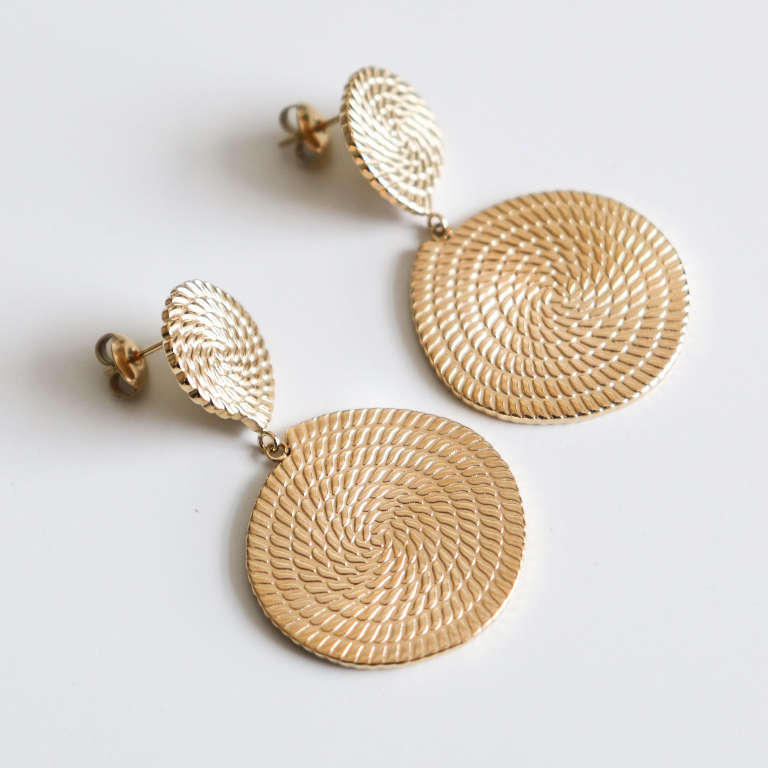 Auskari - Grācija, sieviešu auskari no zelta pārklājuma juveliertērauda ar apļveida, teksturētu spirāles rakstu uz kniedēm un lielākām ripām, skaisti izcelti uz balta fona.