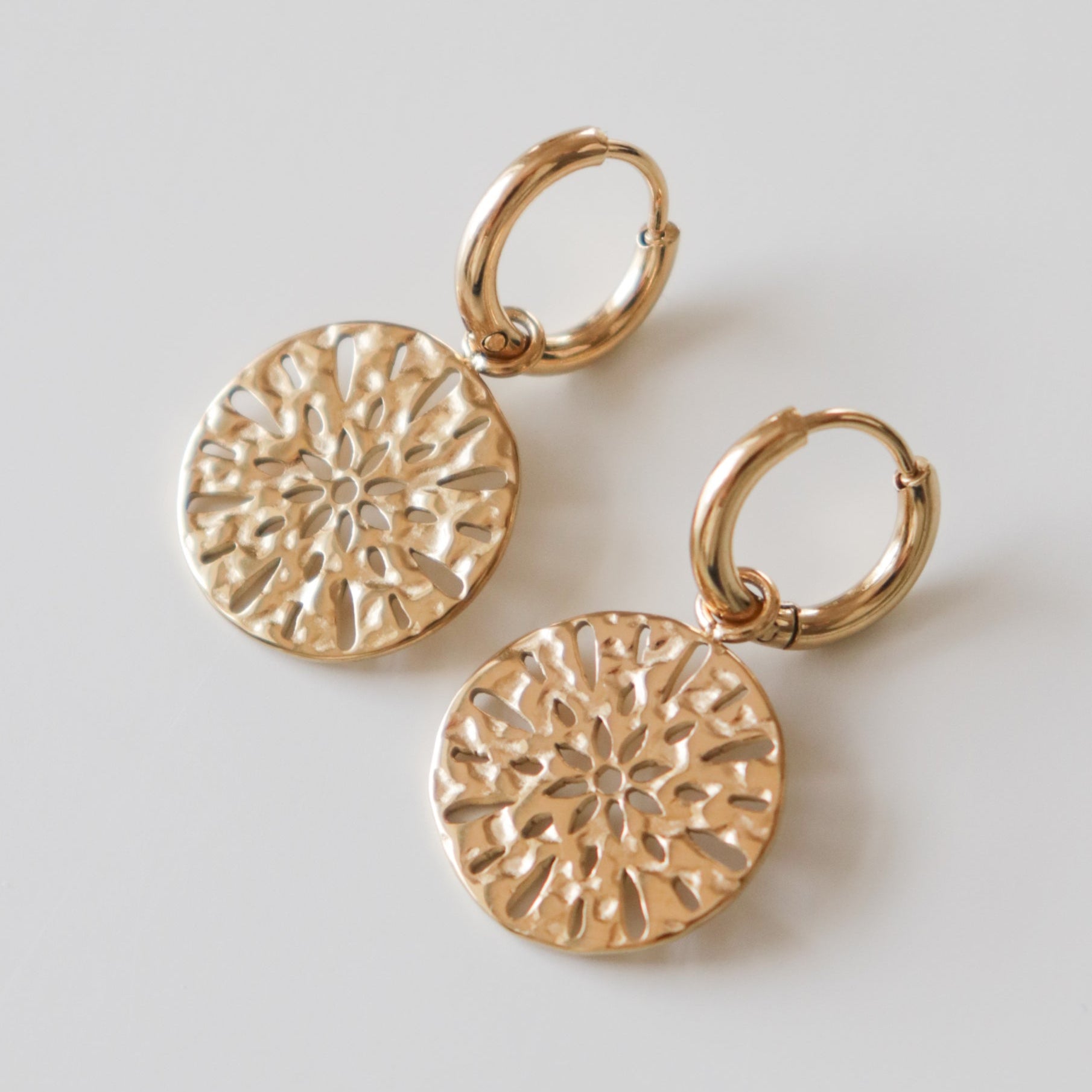 Como Jewelry "Auskari - Saules raksts": mūsdienīgi juveliertērauda auskari ar zelta aplīšiem un apaļiem, teksturētiem piekariņiem ar izgrieztu saules motīvu, izvietoti uz balta fona.