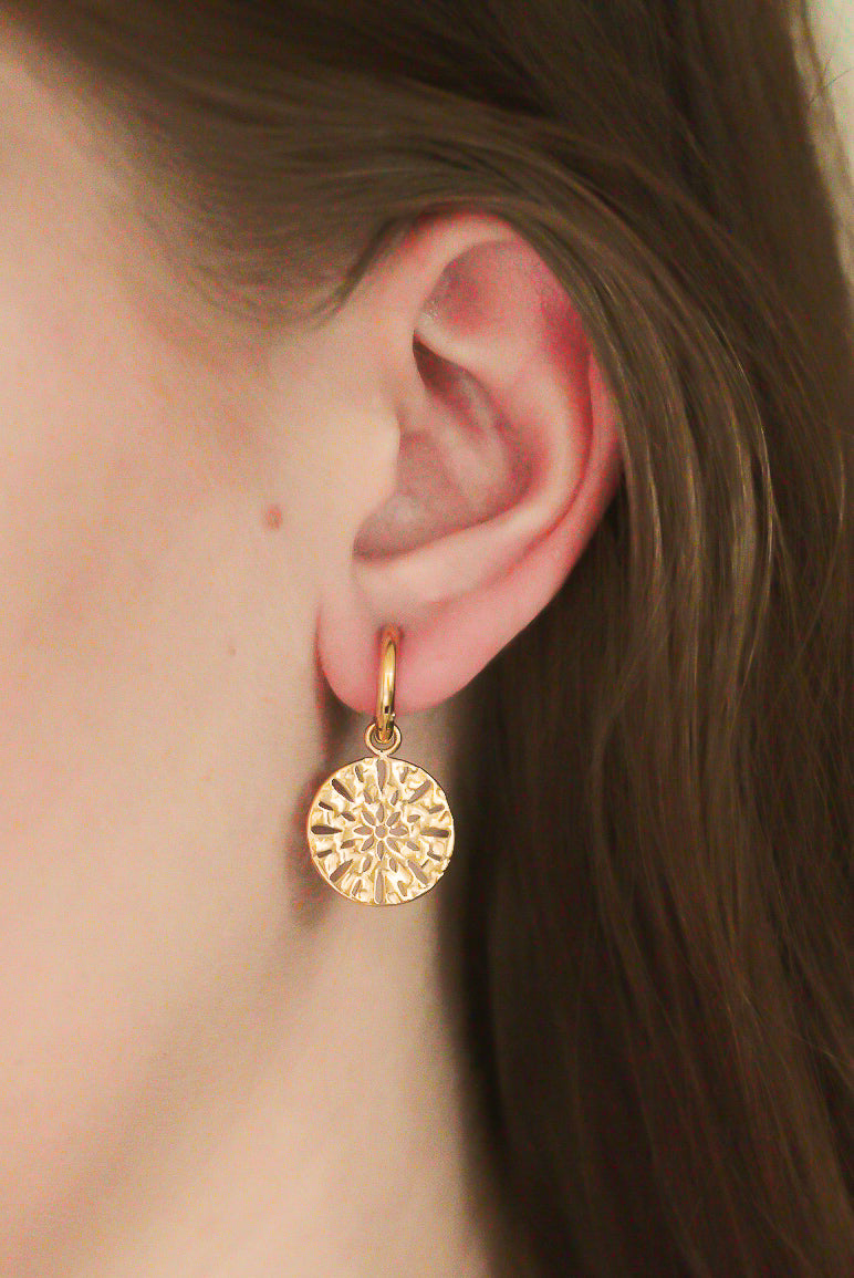 Tuvplāns, kurā redzama persona ar Como Jewelry "Auskari - Saules raksts" - teksturētiem, apaļiem, apaļiem, zeltītiem auskariem ar saules plankumainiem uz maziem zelta riņķīšiem. Mūsdienīgi un ideāli piemēroti ikdienas nēsāšanai, un personas brūnie mati ir aizbīdīti aiz auss.