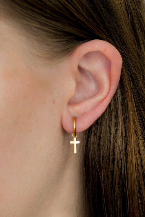 Tuvplāns uz cilvēka auss, kurā valkā Como Jewelry Auskari - ar krustu, nelielu zelta riņķa auskaru ar zelta krusta piekariņu. Redzami brūnie mati; fons nav fokusēts.