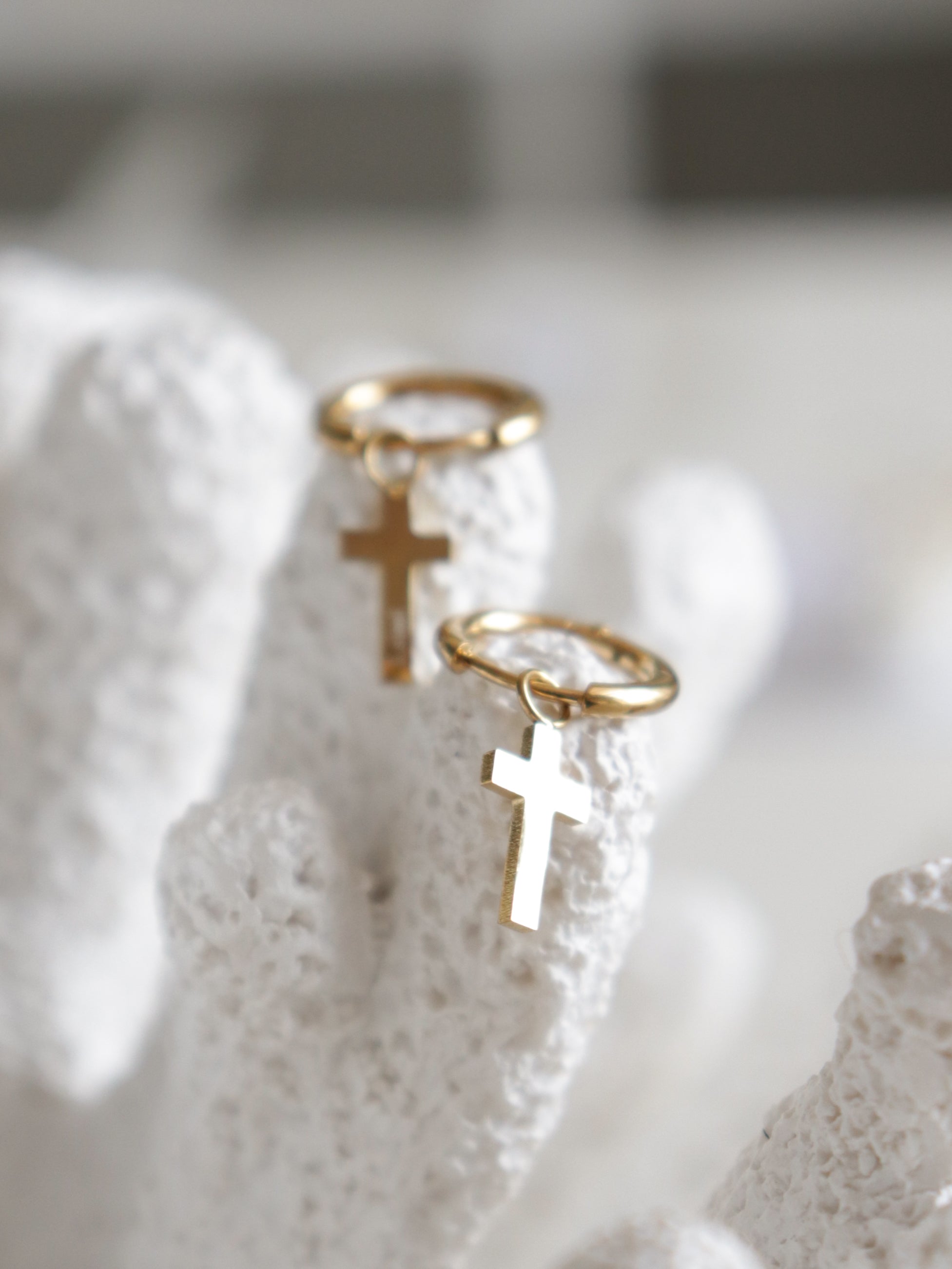 Como Jewelry Auskari - ar krustu: zelta riņķi ar krusta piekariņiem, kas attēloti uz koraļļiem līdzīgas tekstūras baltas virsmas. Maigi izplūdušais fons izceļ šos modernos auskarus ikdienas valkāšanai.