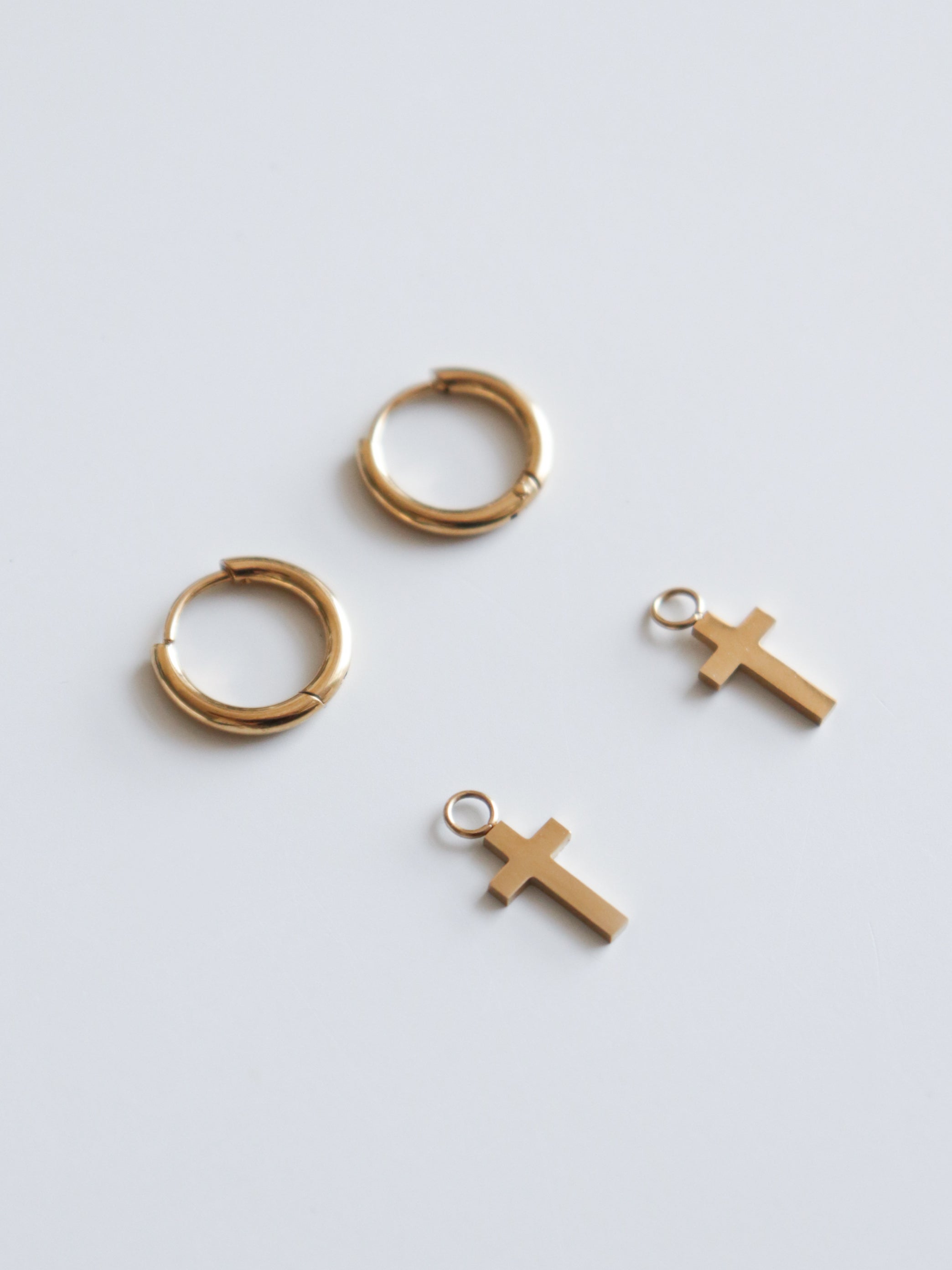 Divi Como Jewelry zelta riņķveida auskari "Auskari - ar krustu" ar diviem maziem zelta krustiņu piekariņiem uz vienkrāsainas baltas virsmas.