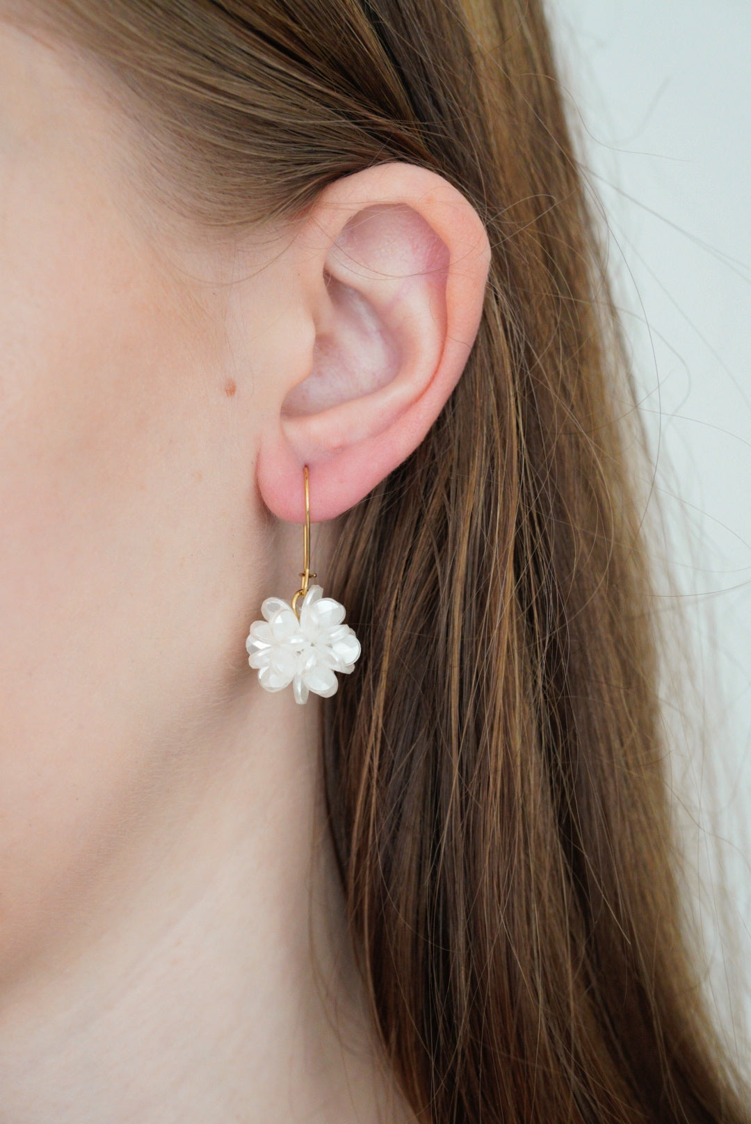 Detalizēts tuvplāns sievietes ausij, kurai ir Como Jewelry "Auskari - Rītausma" zelta āķīšu auskari ar baltu ziedu kopuma rakstu; brūnie mati ir aizbīdīti aiz auss, uz neitrāla fona.