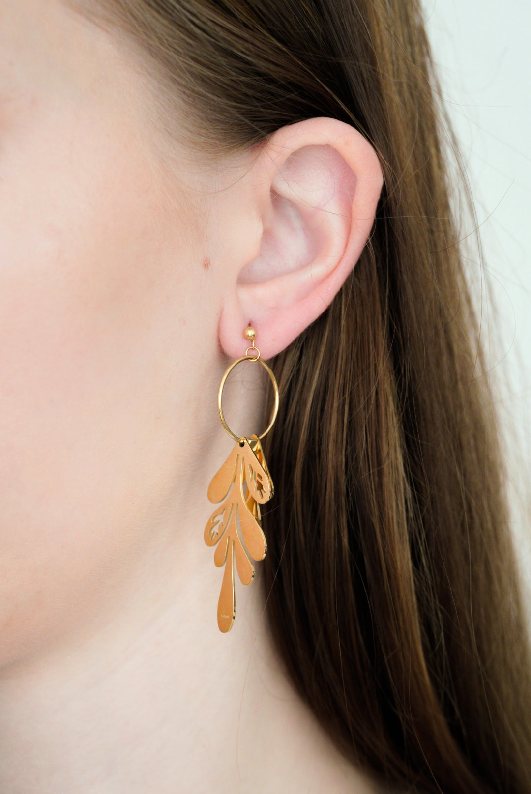 Tuvplāns sievietes ausij ar gariem brūniem matiem, kurai ir Como Jewelry Auskari - Brīze, eleganti lapu formas auskari, kas piestiprināti pie maza riņķīša.