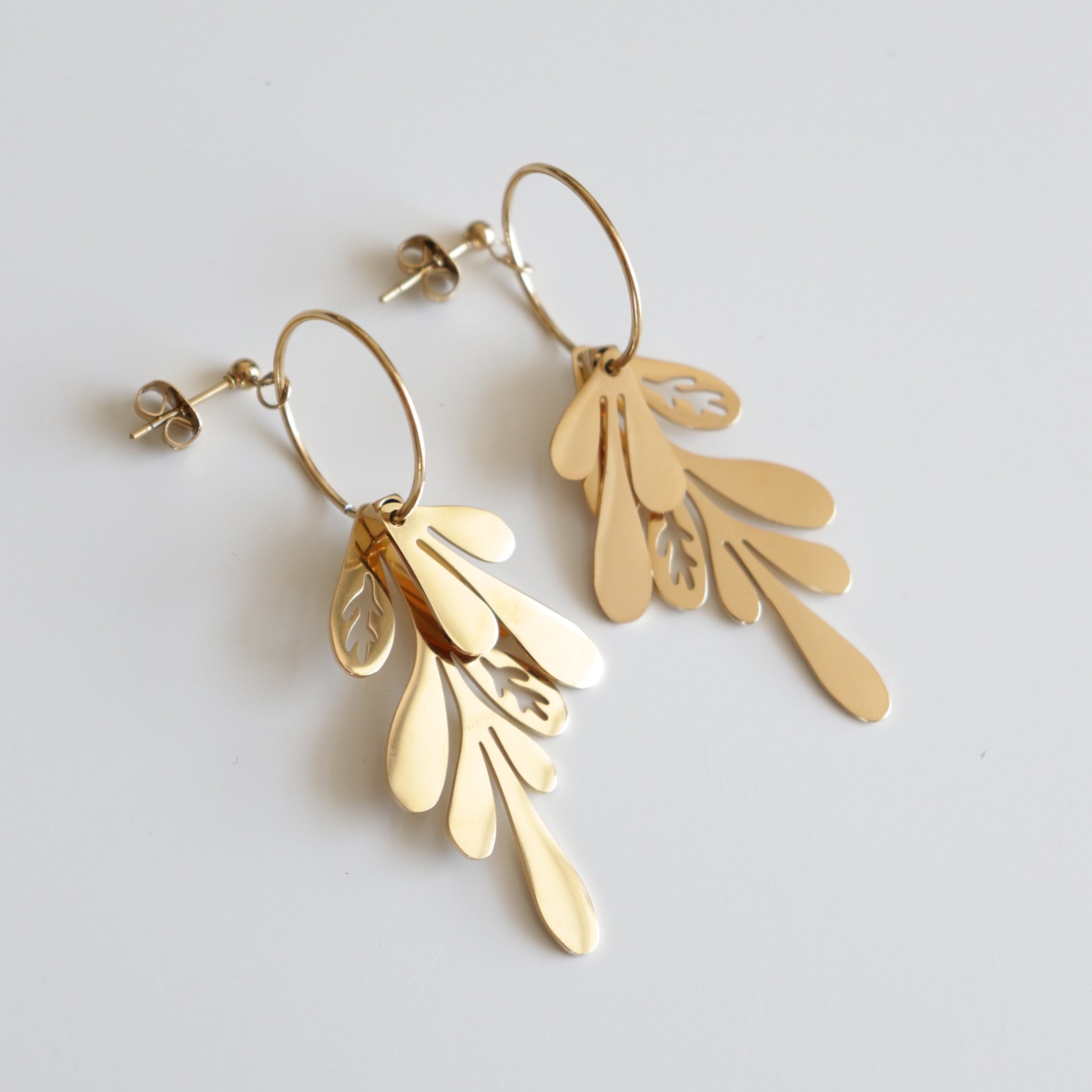 Como Jewelry "Auskari - Brīze" ir juveliertērauds zelta piekaramie auskari ar pārklājošiem asaru un lapu formas piekariņiem uz apaļiem riņķiem, kas attēloti uz balta fona.