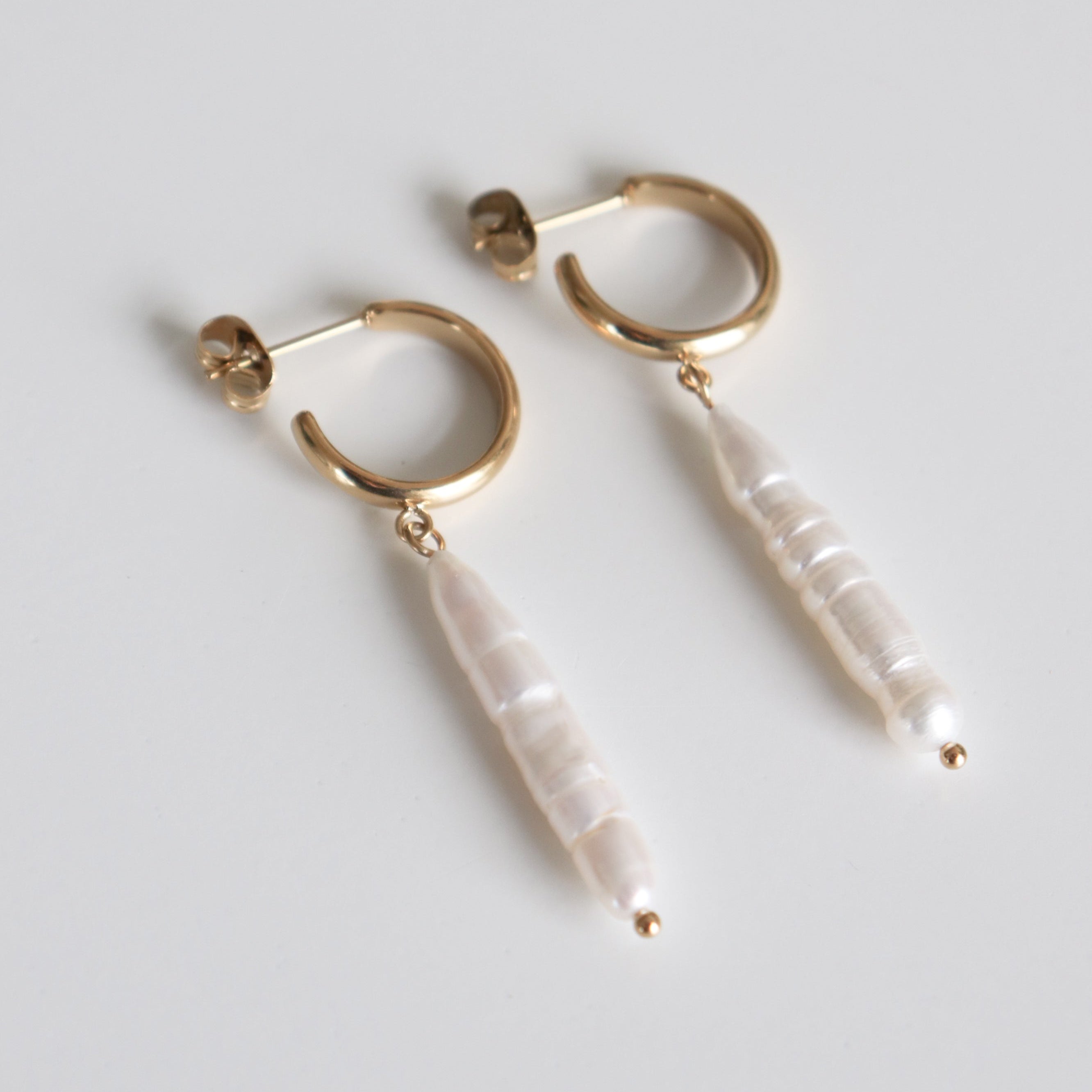 Como Jewelry "Auskari - Lagūnas pērles" ir ar iegarenām, neregulārām baltām pērlēm, kas karājas no katra riņķa un ir uz vienkārša gaiši pelēka fona.