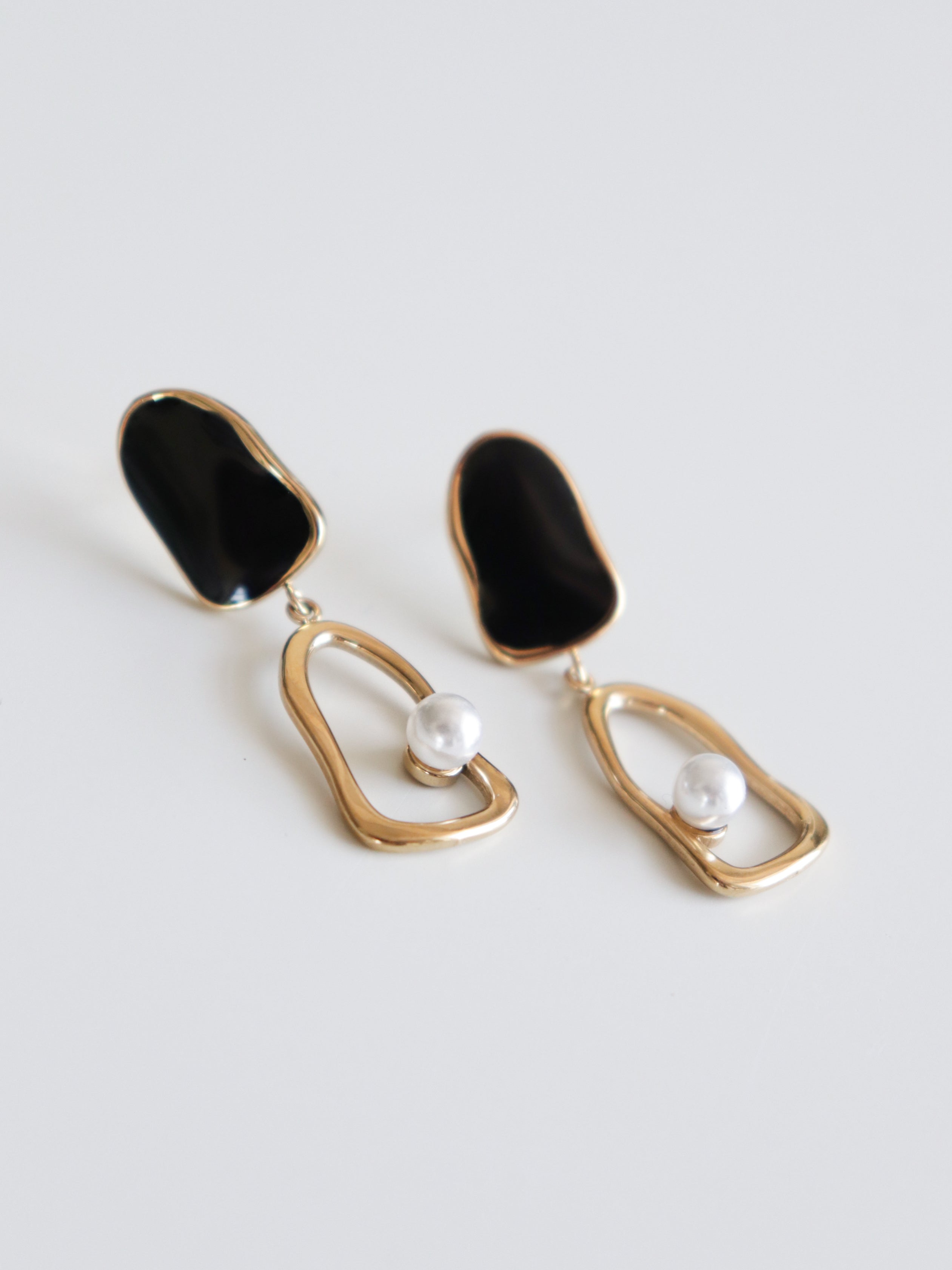 Como Jewelry "Auskari - Atspulgs" ir asimetriski auskari ar melnu emalju, neregulārām zelta cilpiņām un atsevišķām baltām pērlēm. Šie ūdensizturīgi izstrādājumi ir attēloti uz vienkārša balta fona.