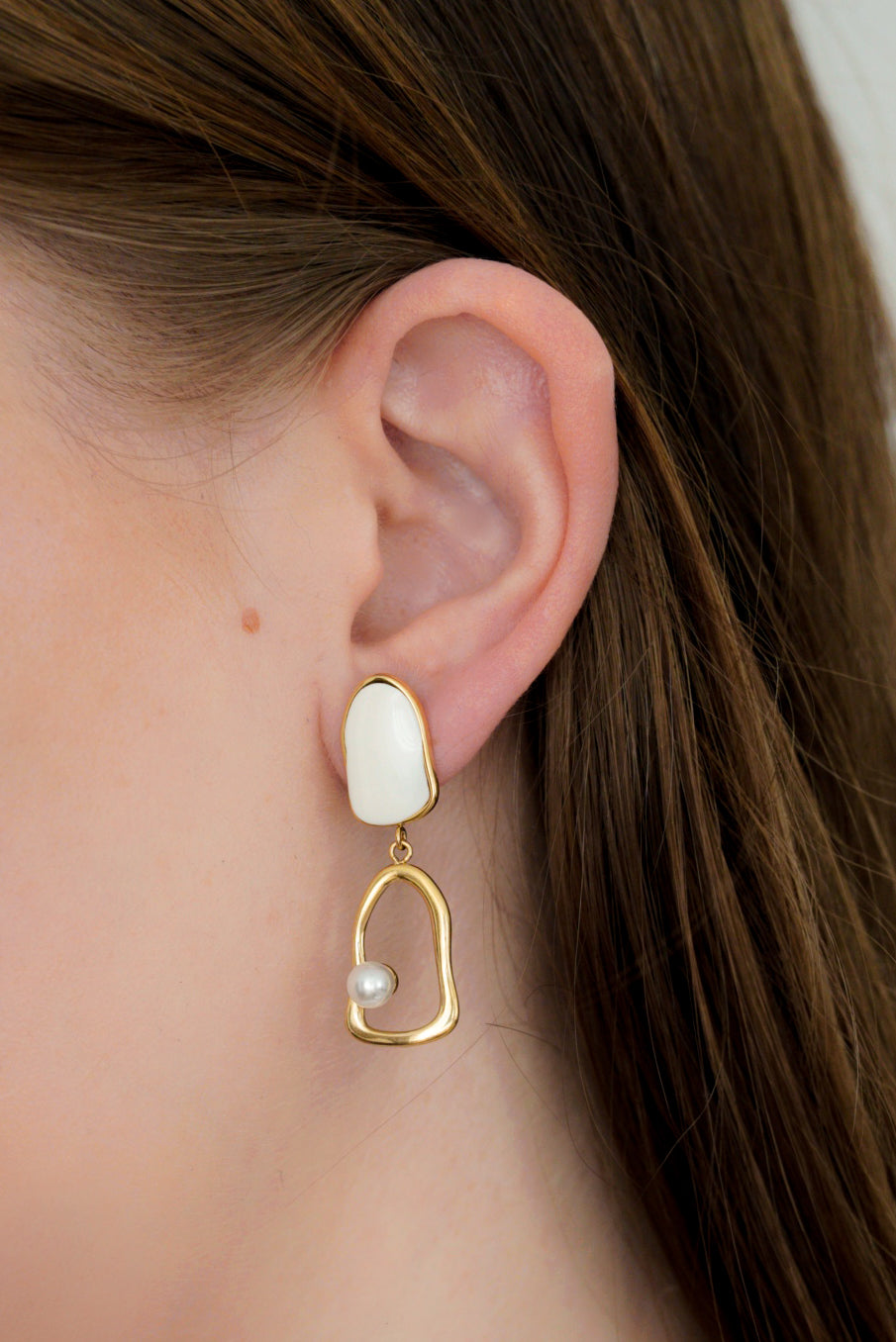 Tuvplāns sievietes ausij, kurai ir Como Jewelry Auskari - Atspulgs: moderni, zelta krāsas, ūdensizturīgi auskari ar abstraktu juveliertērauda ietvaru, baltu akmentiņu un vienu pērles akcentu.